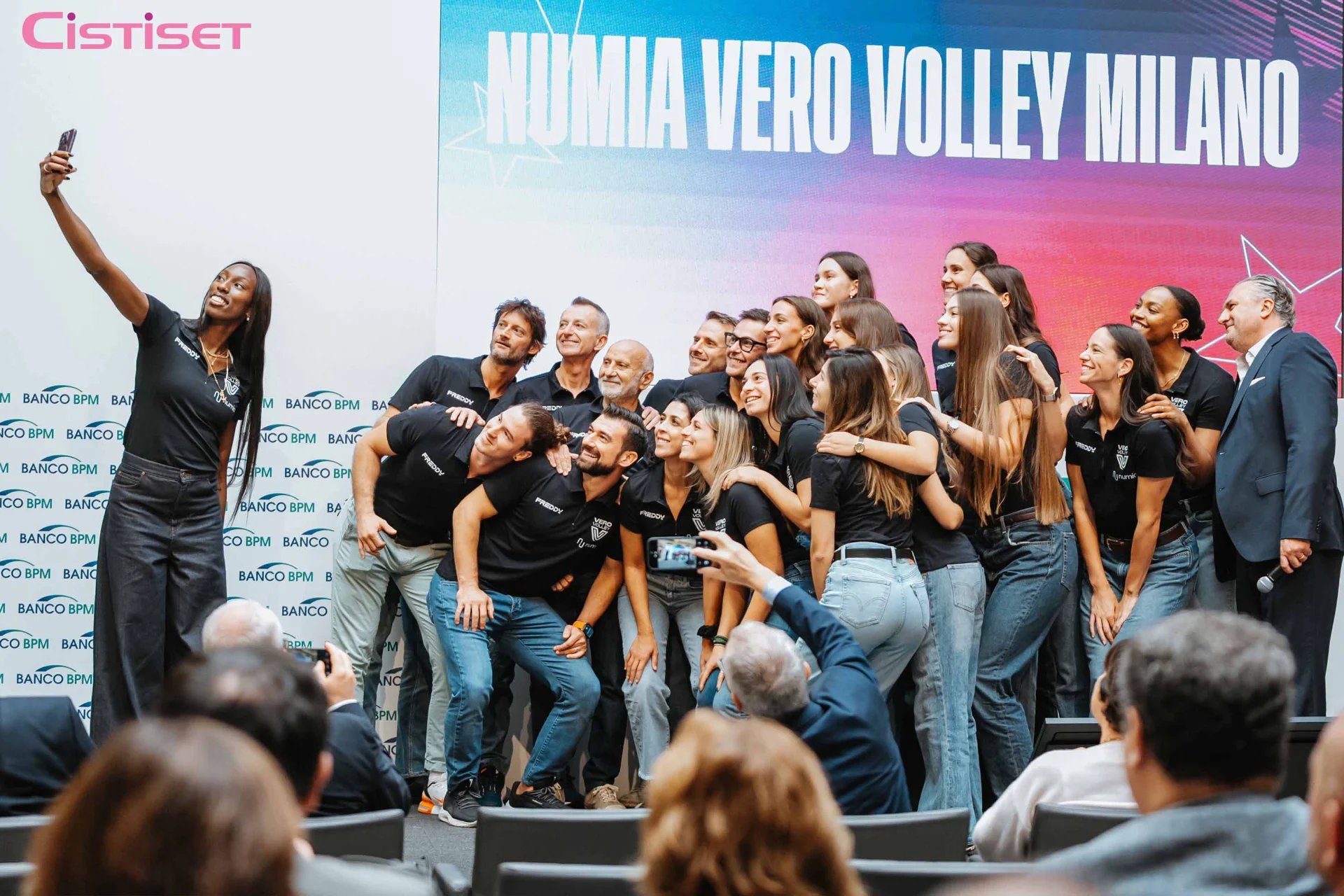 Giocatrici di pallavolo e staff Vero Volley Milano sorridono mentre una giocatrice scatta un selfie con lo smartphone.